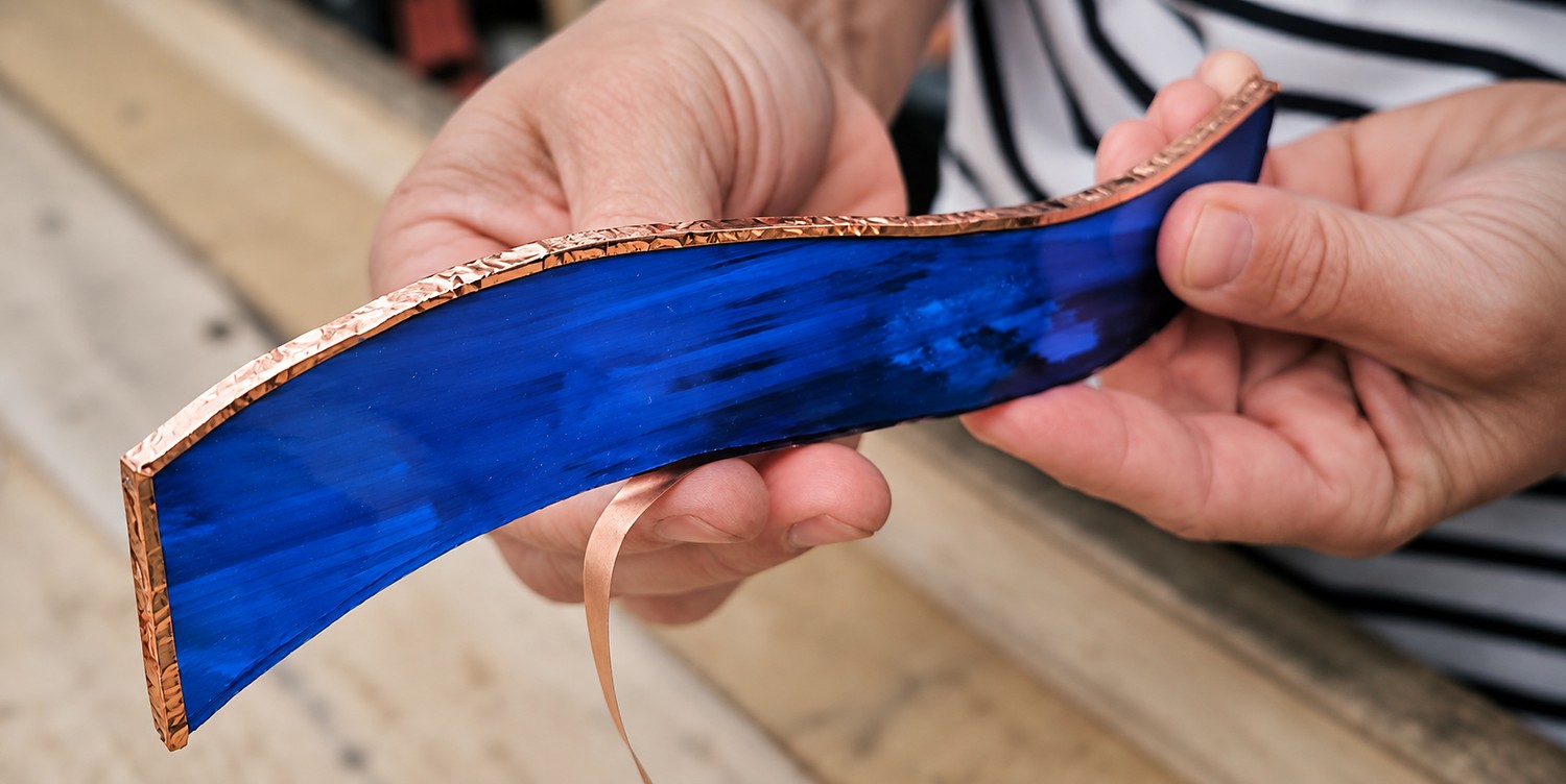 Pose manuelle d’un ruban de cuivre autour d’une pièce de verre bleu selon la technique Tiffany
